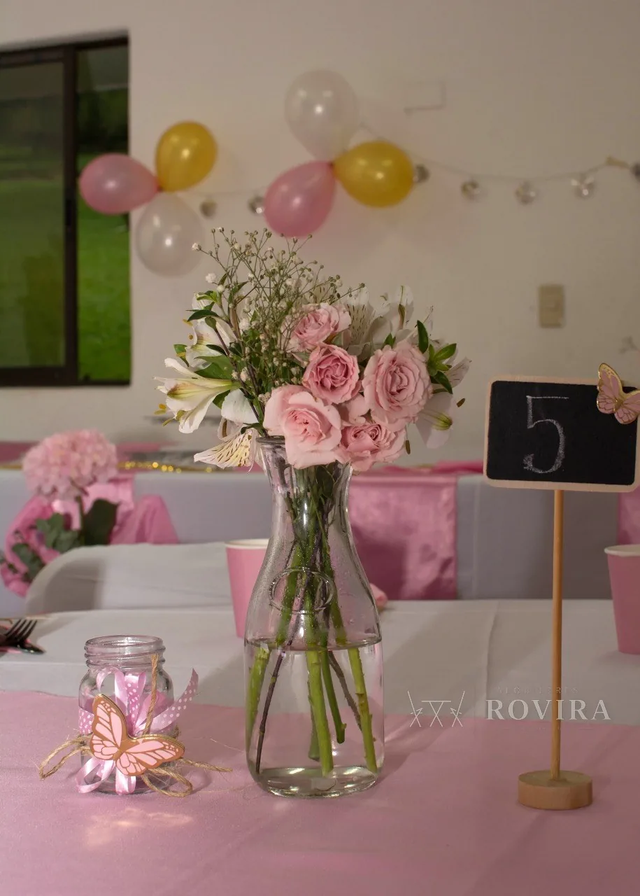 Montaje de baby shower en rosa: centro de flores, mariposas y mesas vestidas en tonos pastel, con número de mesa en pizarra