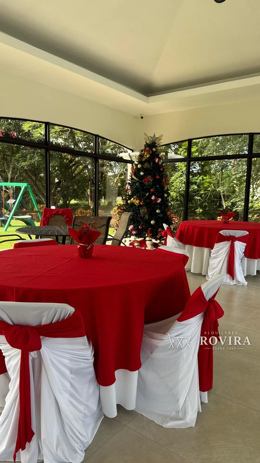 Mesas redondas vestidas en rojo con forro de silla blanco y lazo rojo, salón con árbol de Navidad
