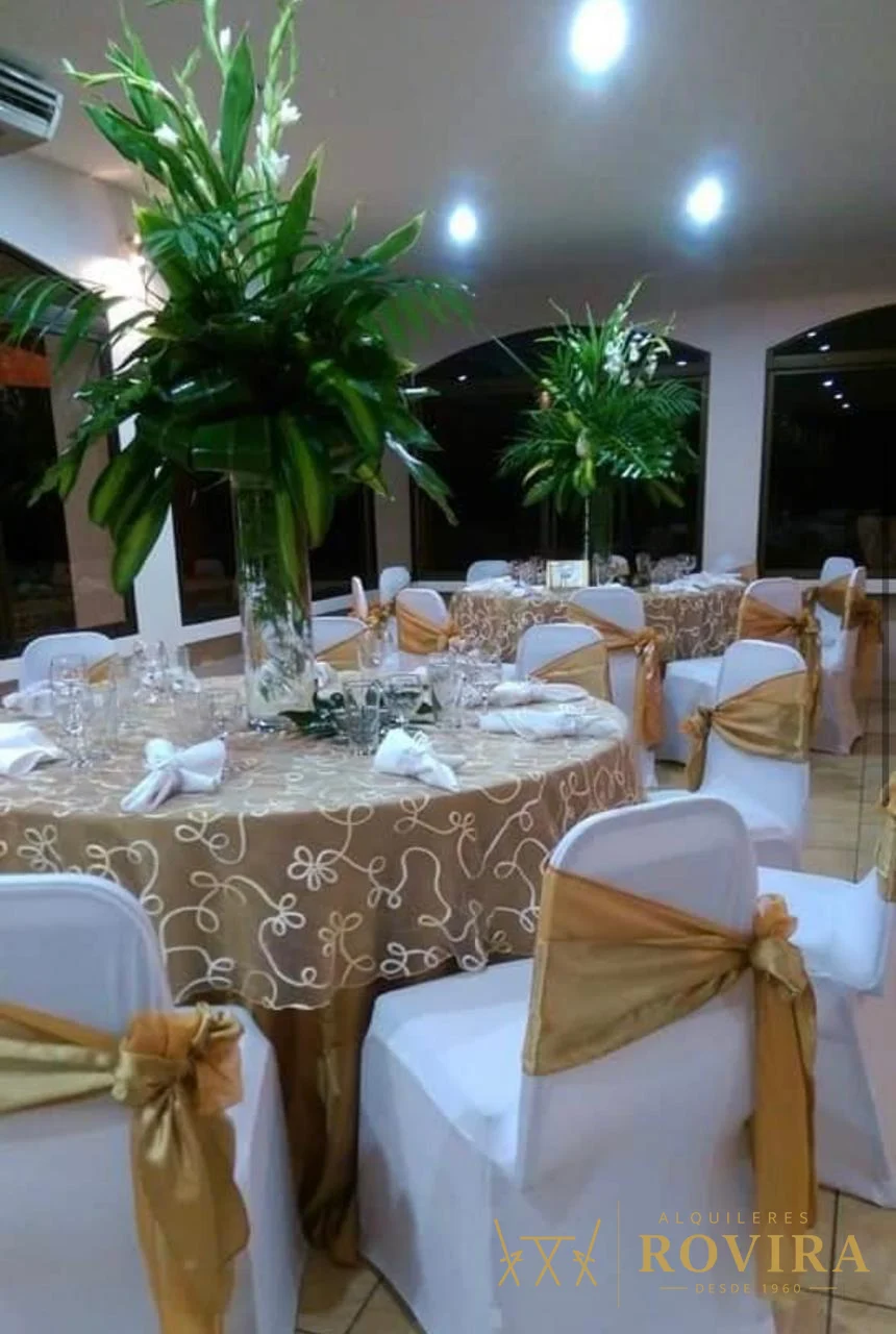 Mesas con mantel dorado champán y forros de silla blancos con lazo dorado, centros verdes altos