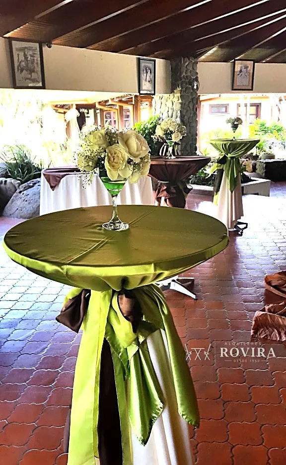 Mesa cocktelera alta y angosta, forrada en verde oliva con lazo café, centro floral blanco