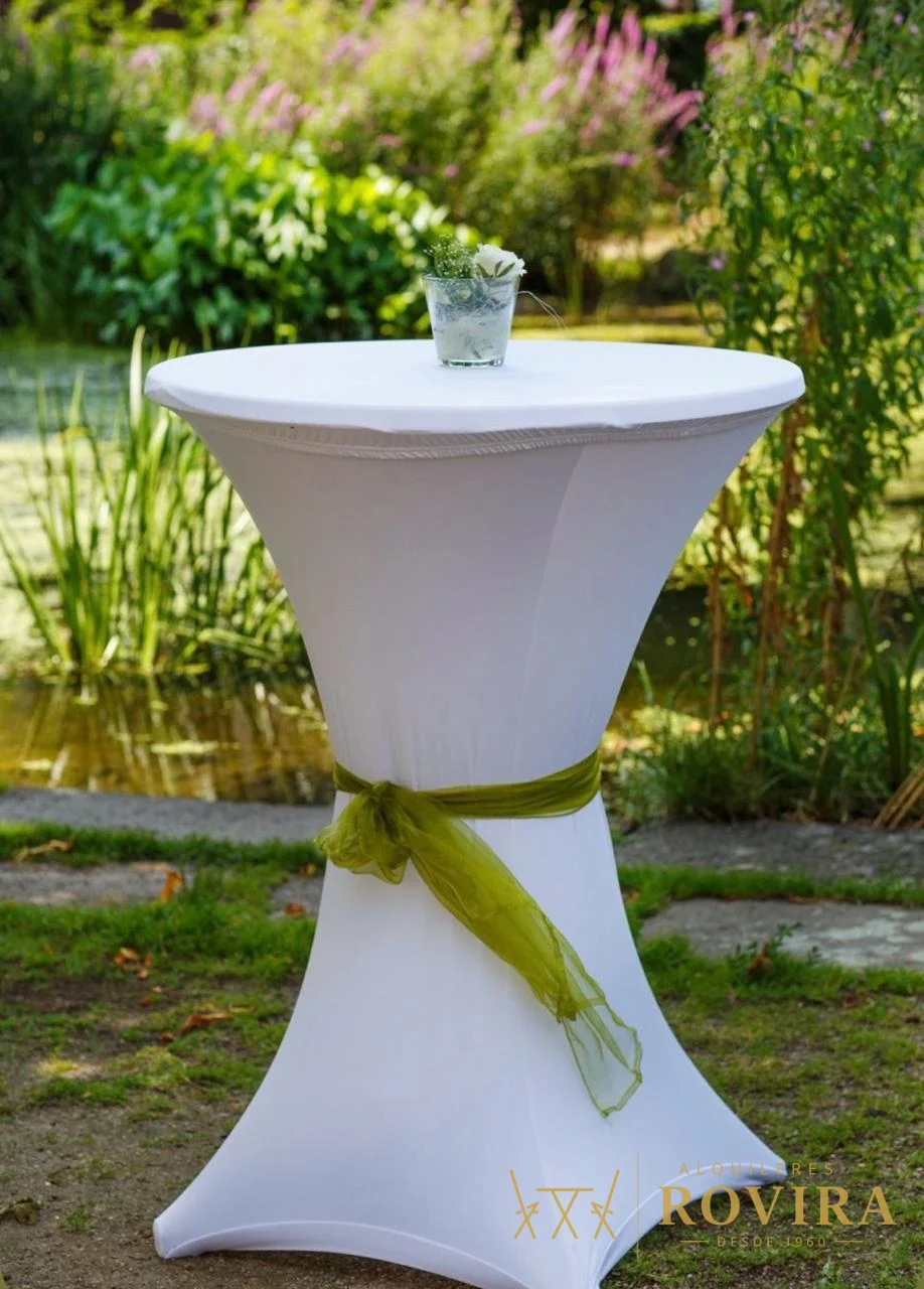 Mesa cocktelera alta y angosta con forro blanco y lazo verde, montada en jardín