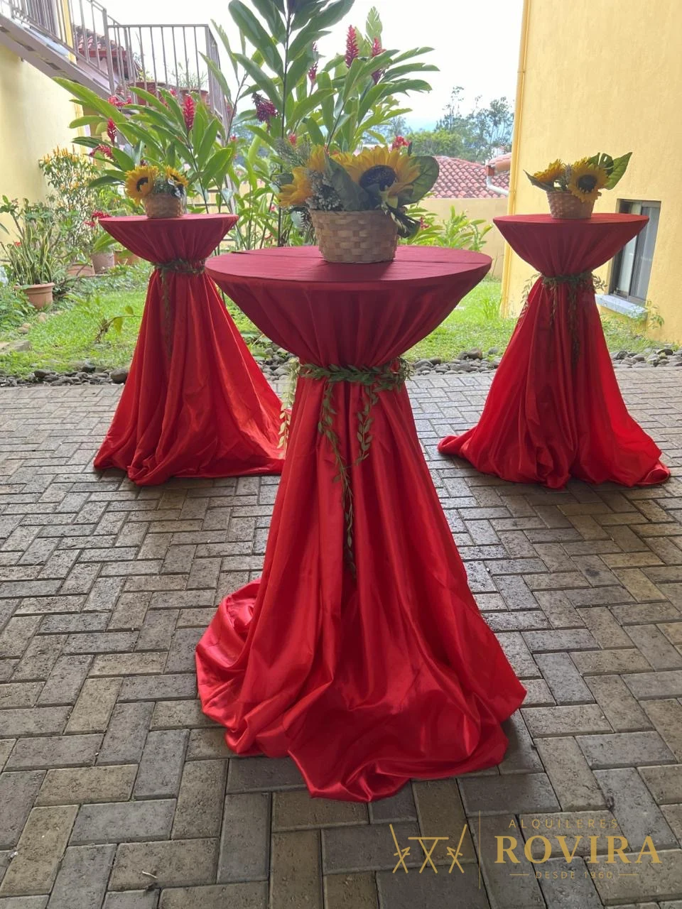 Mesas cocktaileras altas con forro rojo y centros de girasoles, en jardín
