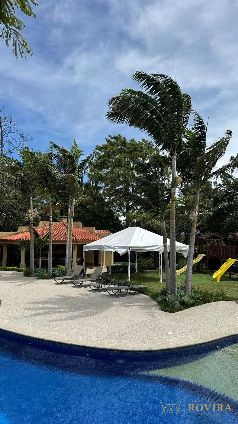 Toldo blanco junto a la piscina de una casa, rodeado de palmeras y jardín, cielo despejado