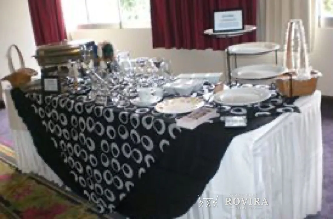 Mesa larga con mantel blanco y negro estampado, montaje de vajilla y cristalería estilo buffet