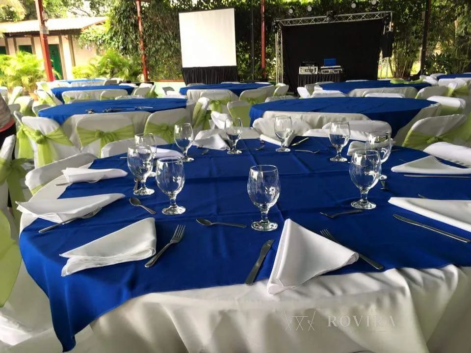 Mesas de evento al aire libre con mantel azul rey y lazos verde limón, copas de vino y agua, servilletas blancas