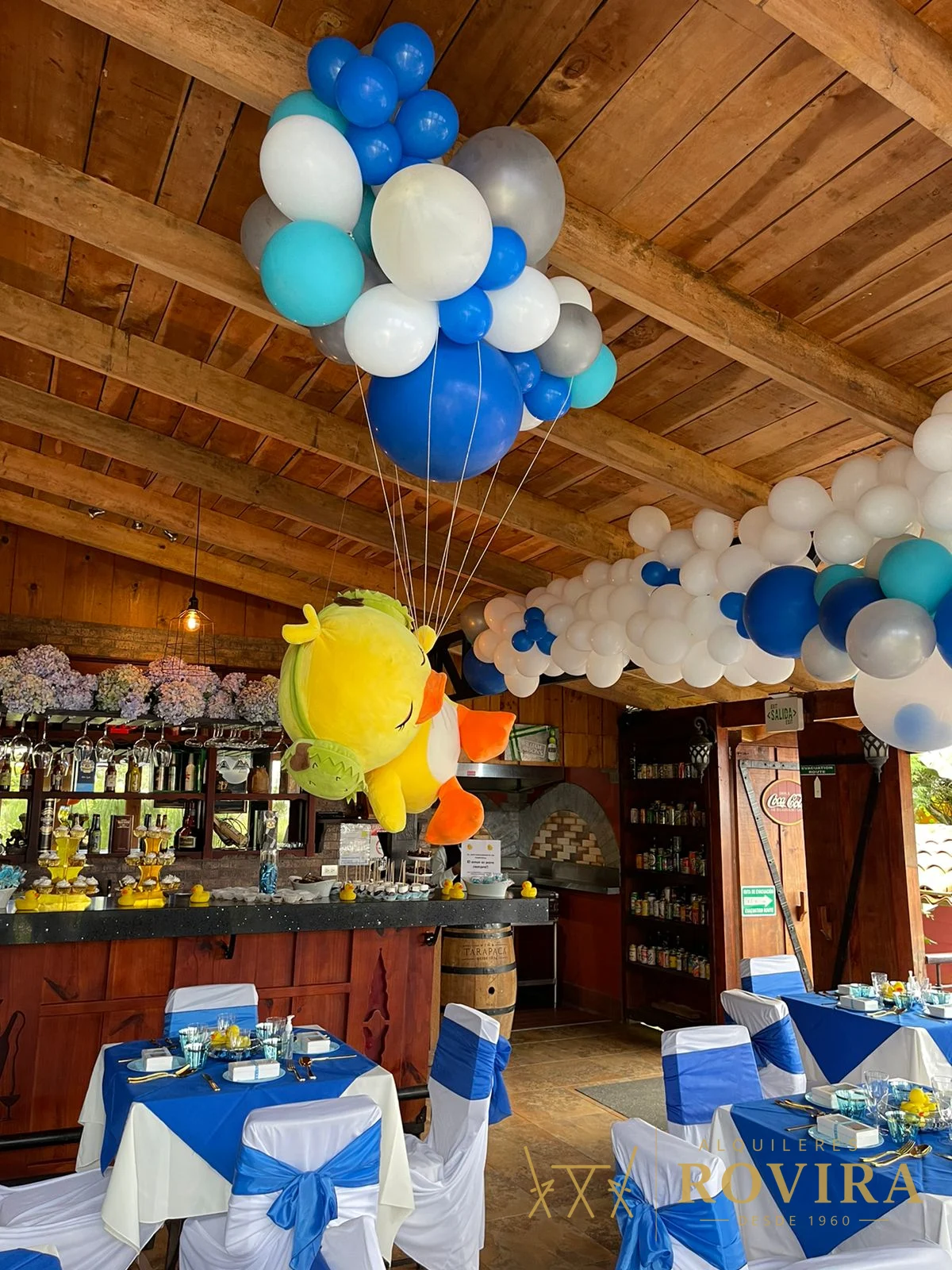 Baby shower de niño con temática de patitos, globos azul y blanco y mesas vestidas en azul, en rancho