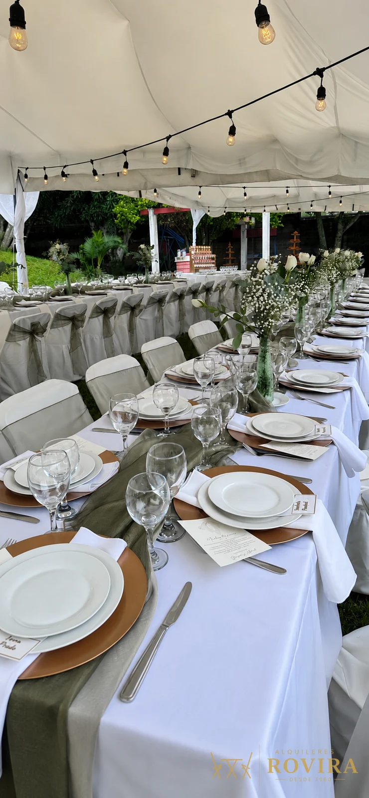 Mesa imperial larga de boda bajo toldo blanco, mantel blanco con camino verde olivo, platos base dorados, cristalería y luces colgantes