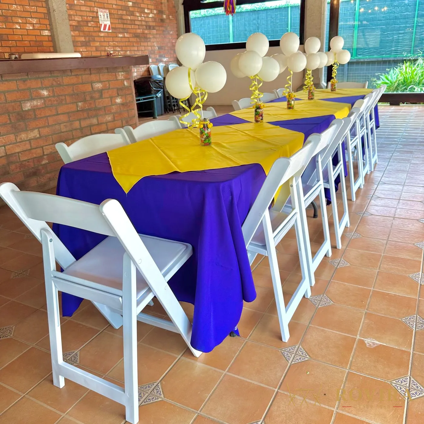 Mesa larga con mantel morado, camino amarillo y sillas plegables blancas acojinadas, con centros de globos blancos