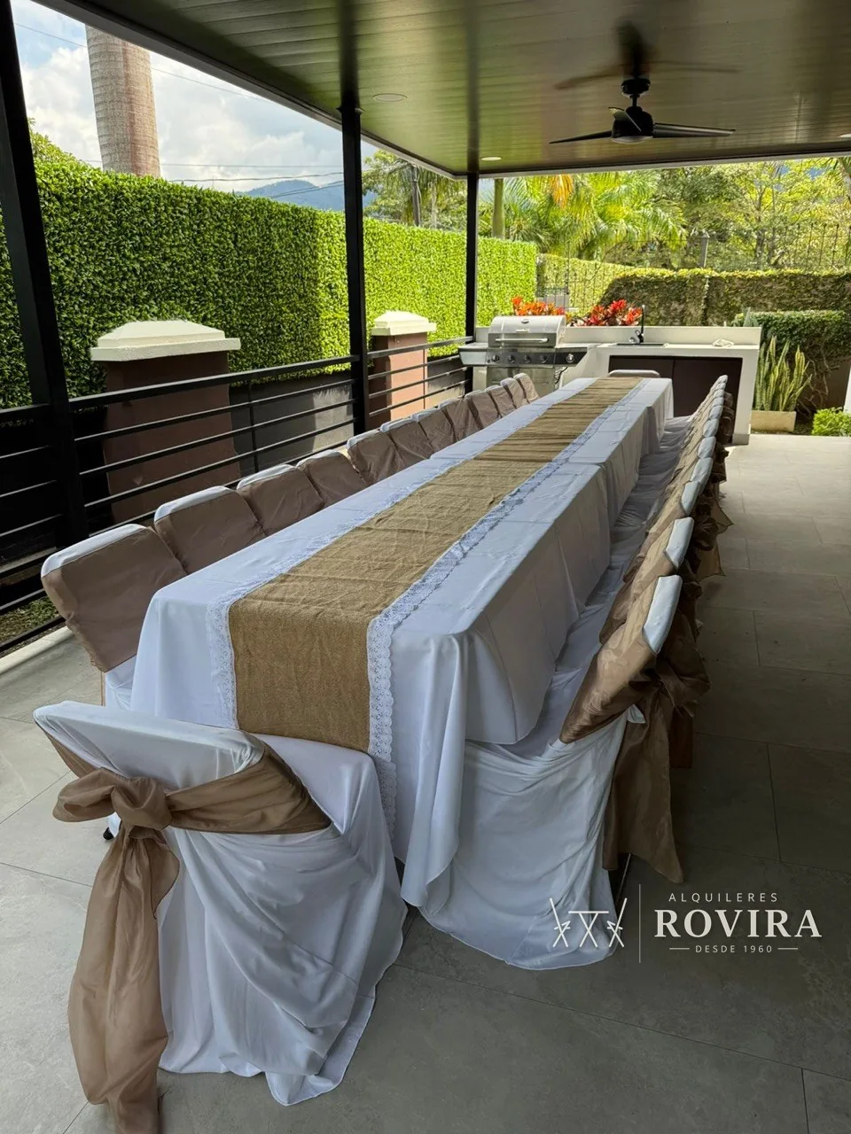 Mesa larga vestida de blanco con camino tipo yute, sillas con lazo café, junto a cocina exterior con parrilla