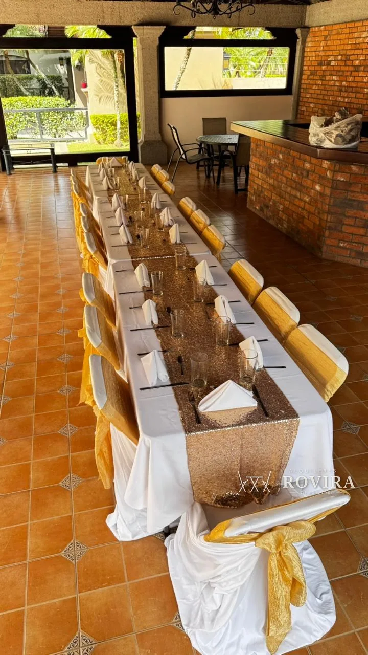 Mesa larga vestida de blanco con camino dorado brillante, sillas con lazo dorado, terraza de piso terracota