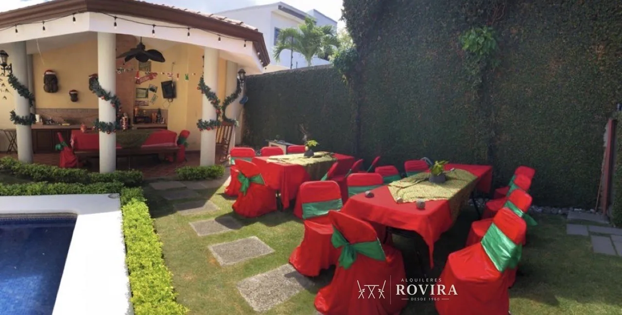 Mesas navideñas vestidas de rojo con lazo verde, junto a piscina y rancho techado decorado para Navidad