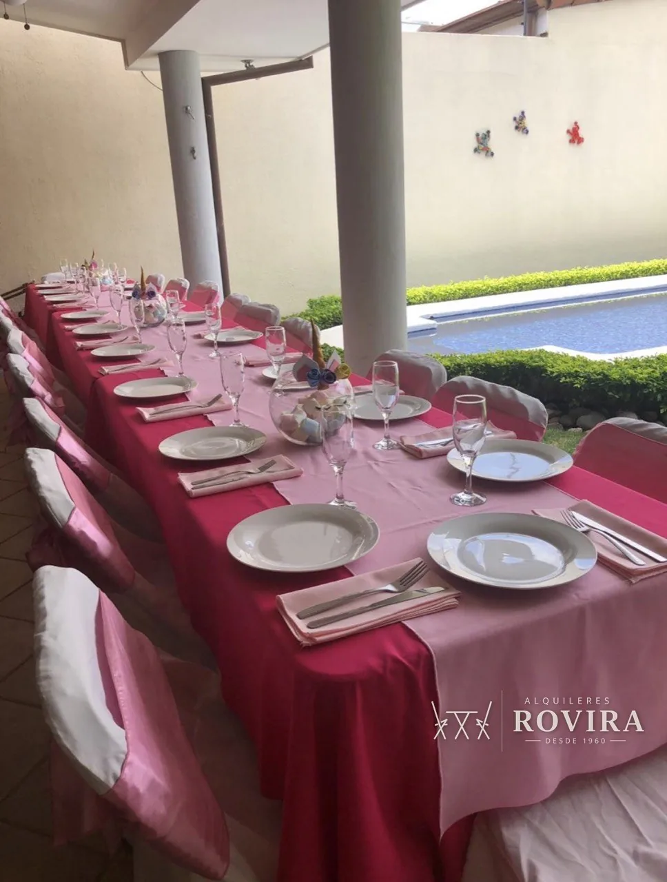 Mesa larga vestida en fucsia y rosa con vajilla blanca y copas, junto a piscina, temática de unicornio