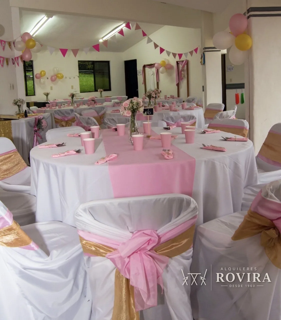Mesas redondas vestidas en blanco con camino rosa y lazo dorado, salón decorado con banderines y globos