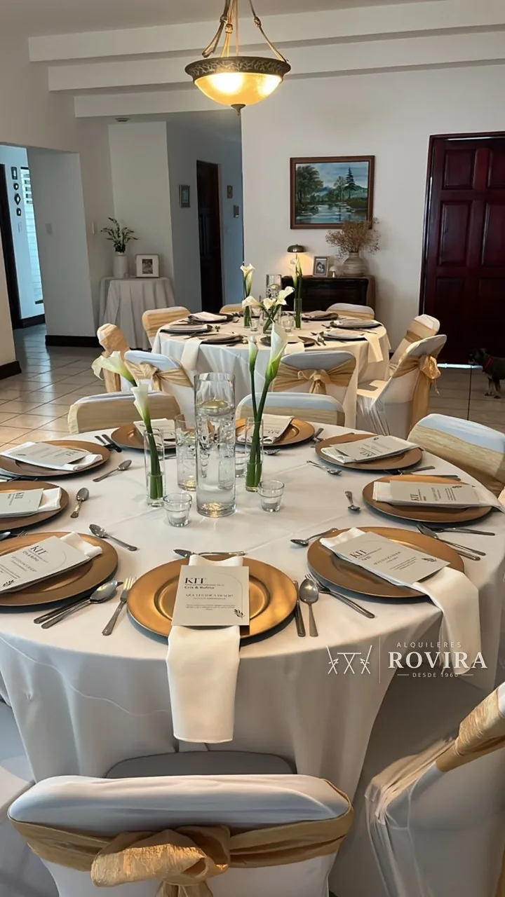 Mesa redonda vestida de blanco con platos base dorados, calas blancas de centro y sillas con lazo dorado, en sala de casa