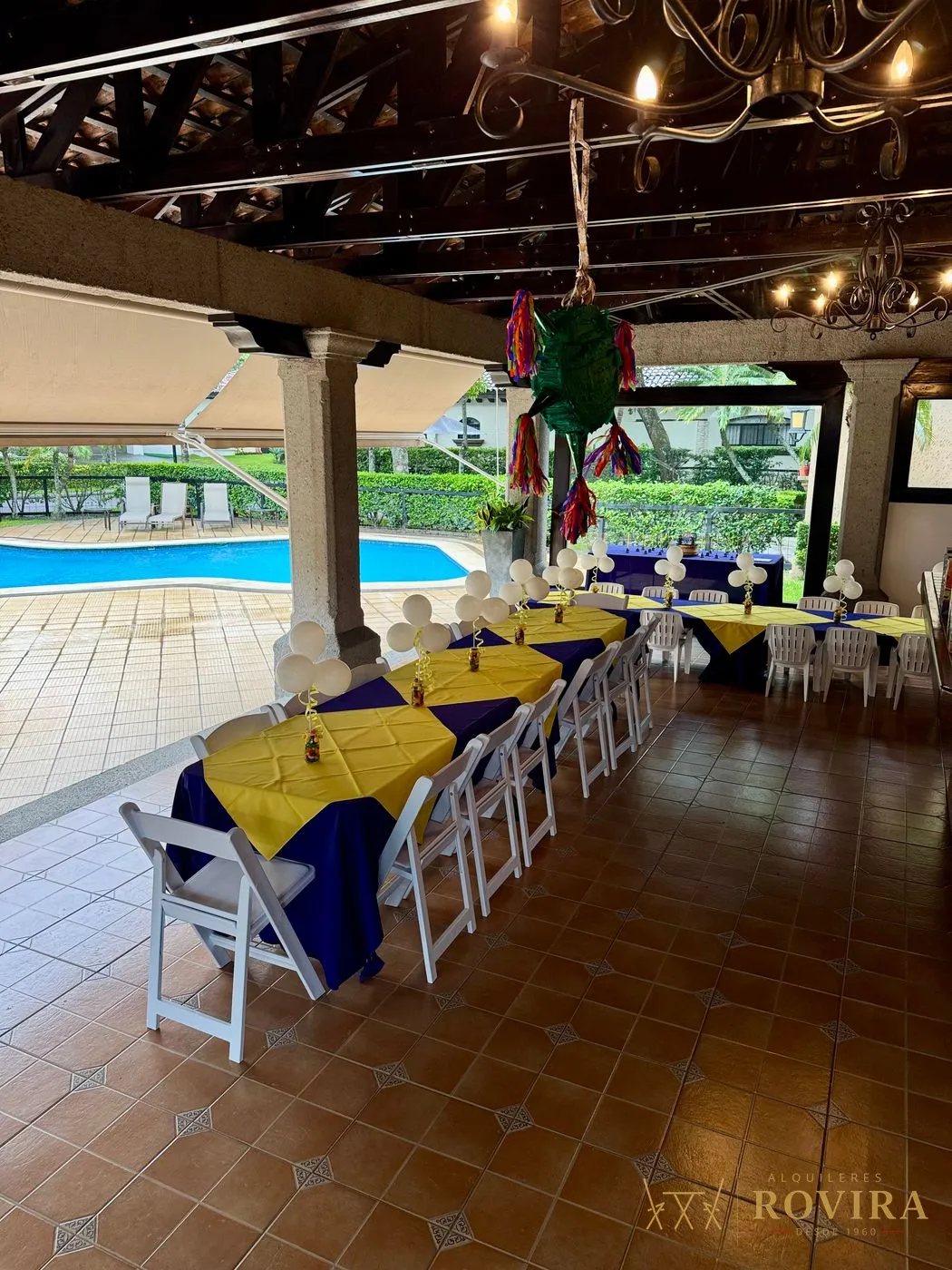 Fiesta con mesas vestidas en morado y amarillo junto a la piscina, bajo rancho con piñata colgante