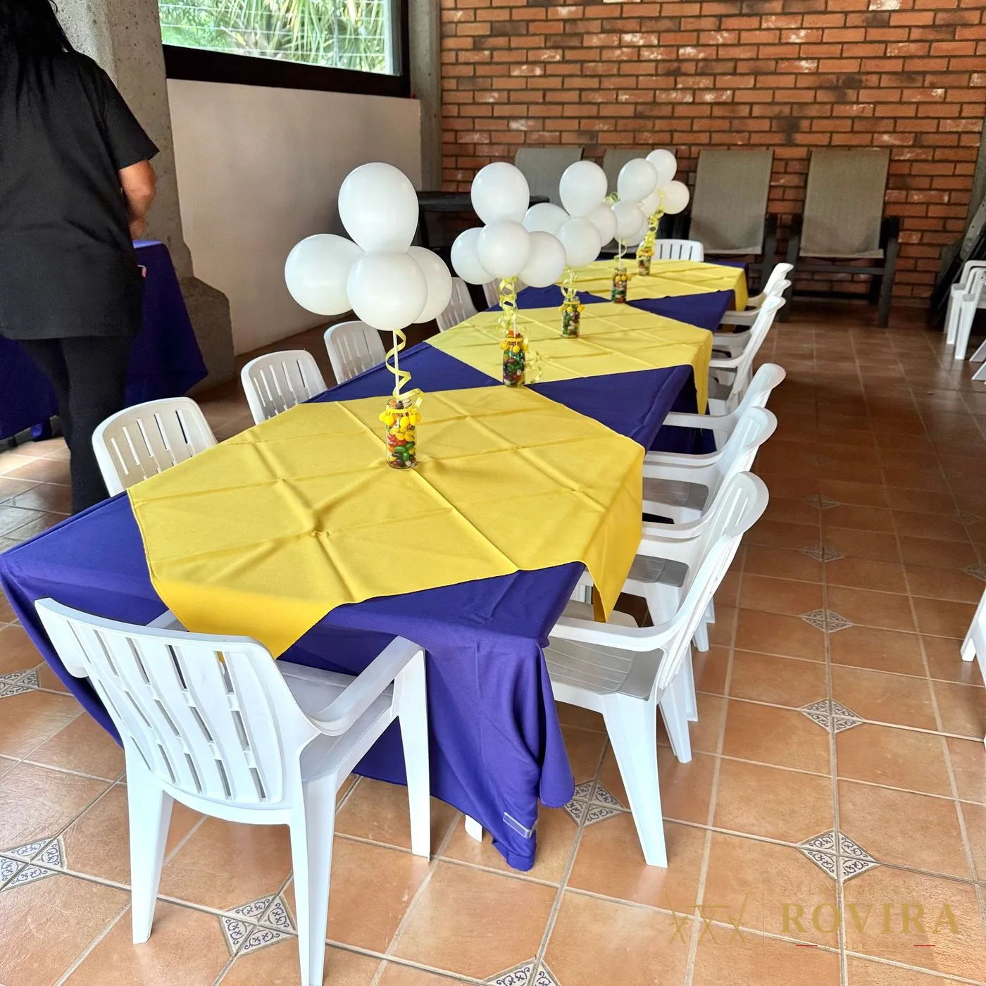 Mesas vestidas en morado y amarillo con centros de globos blancos y frascos de dulces, en terraza