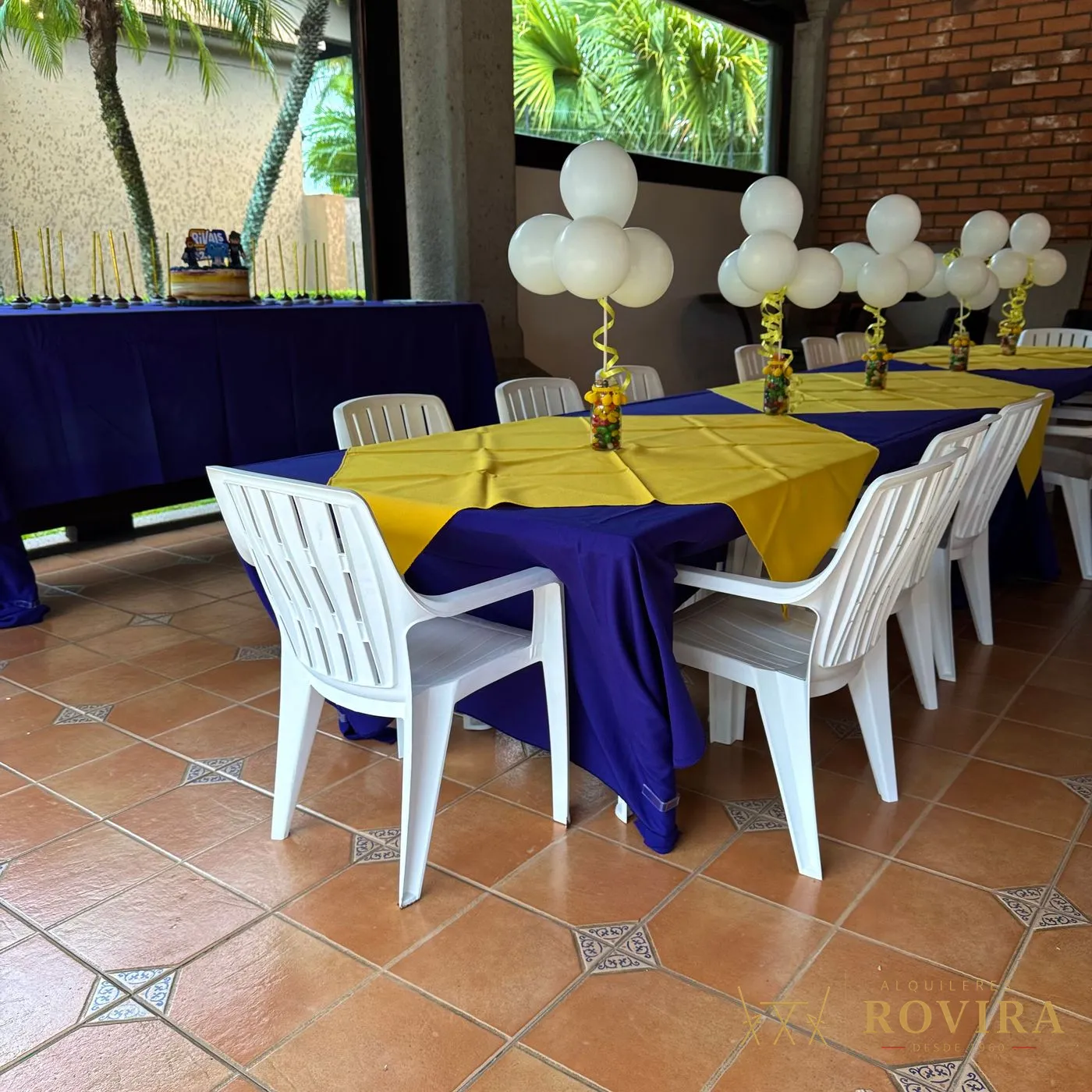 Mesa infantil con mantel morado, camino amarillo y sillas plásticas blancas, con centros de globos