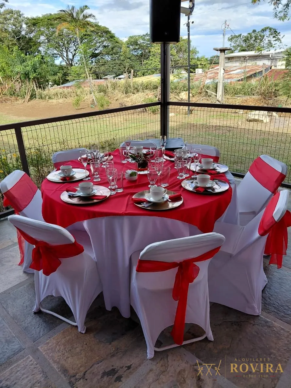 Mesa redonda con mantel rojo sobre blanco, forros de silla con lazo rojo, vajilla y cristalería, en terraza con vista al campo