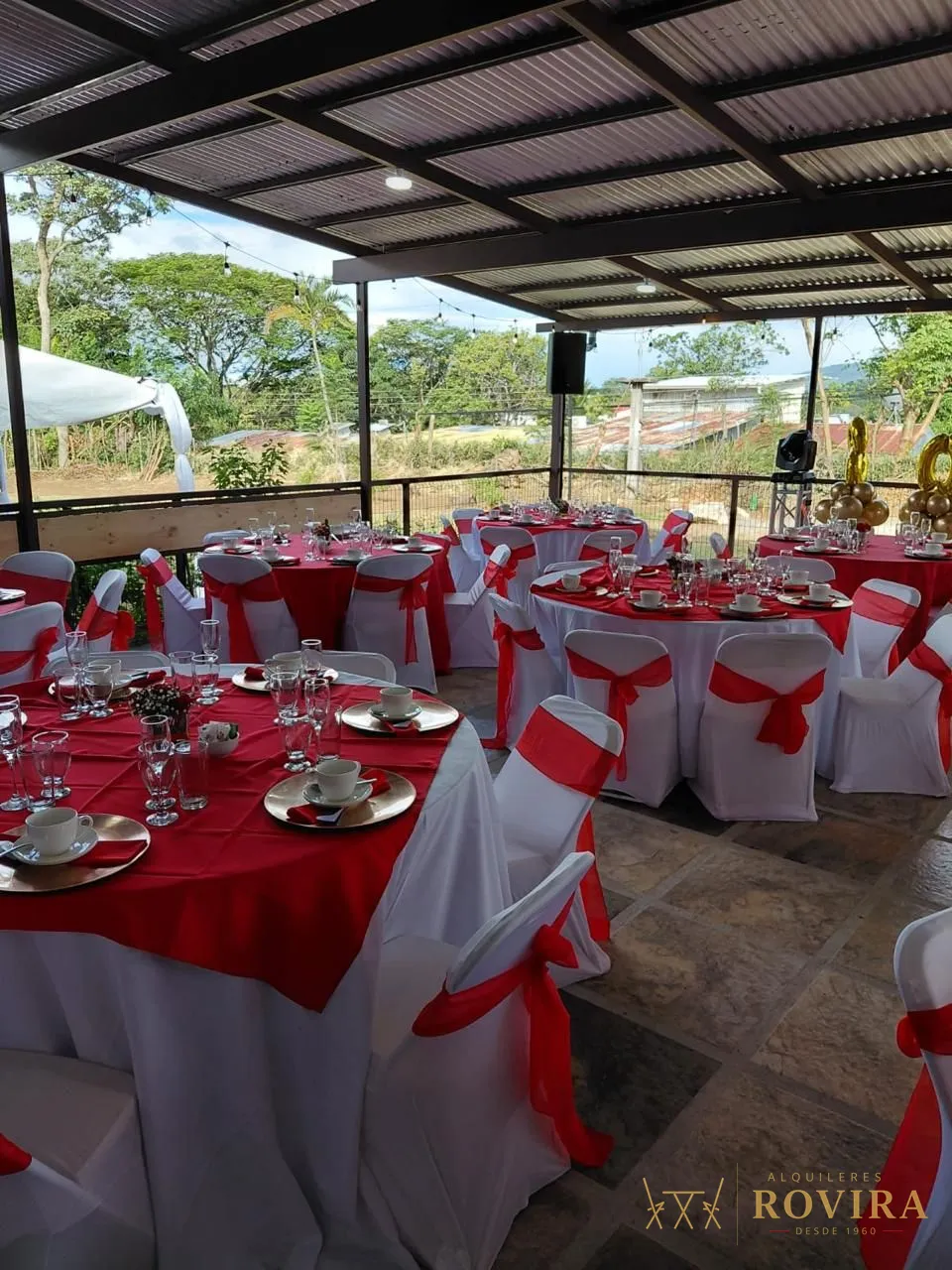 Terraza con varias mesas redondas vestidas en rojo y blanco, forros de silla con lazo rojo y globos dorados de celebración