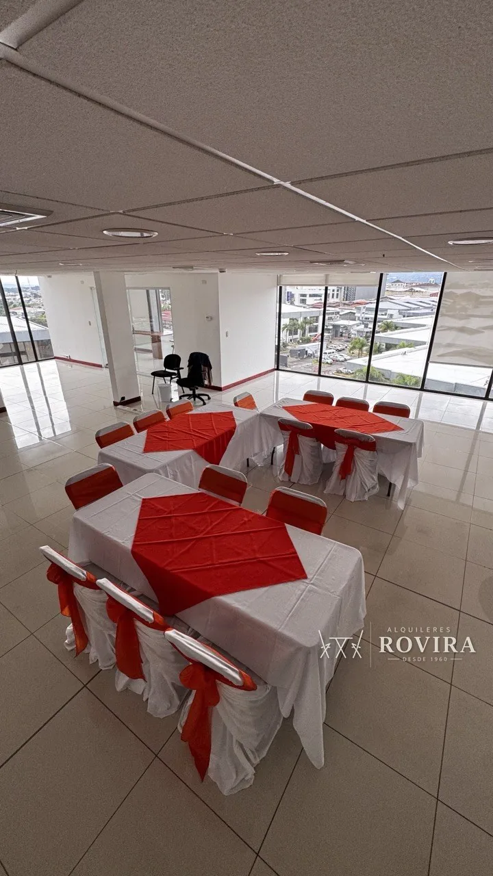 Mesas vestidas en blanco con camino rojo y sillas con lazo rojo, oficina con vista panorámica a la ciudad