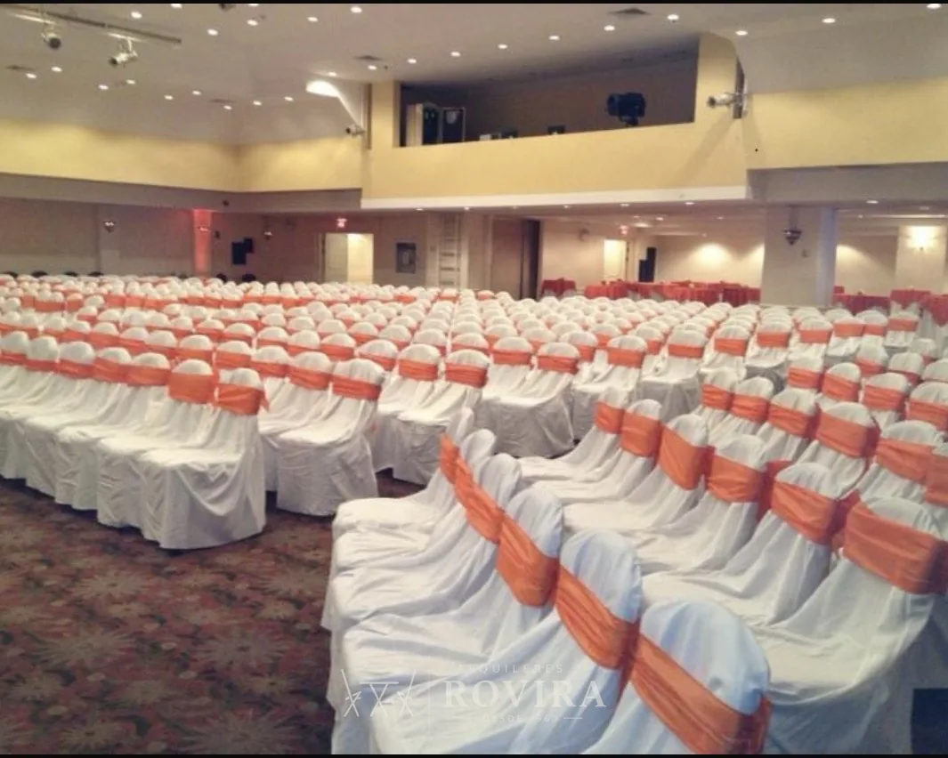 Salón de conferencias montado a gran escala con cientos de sillas forradas blancas y lazos naranja, evento corporativo