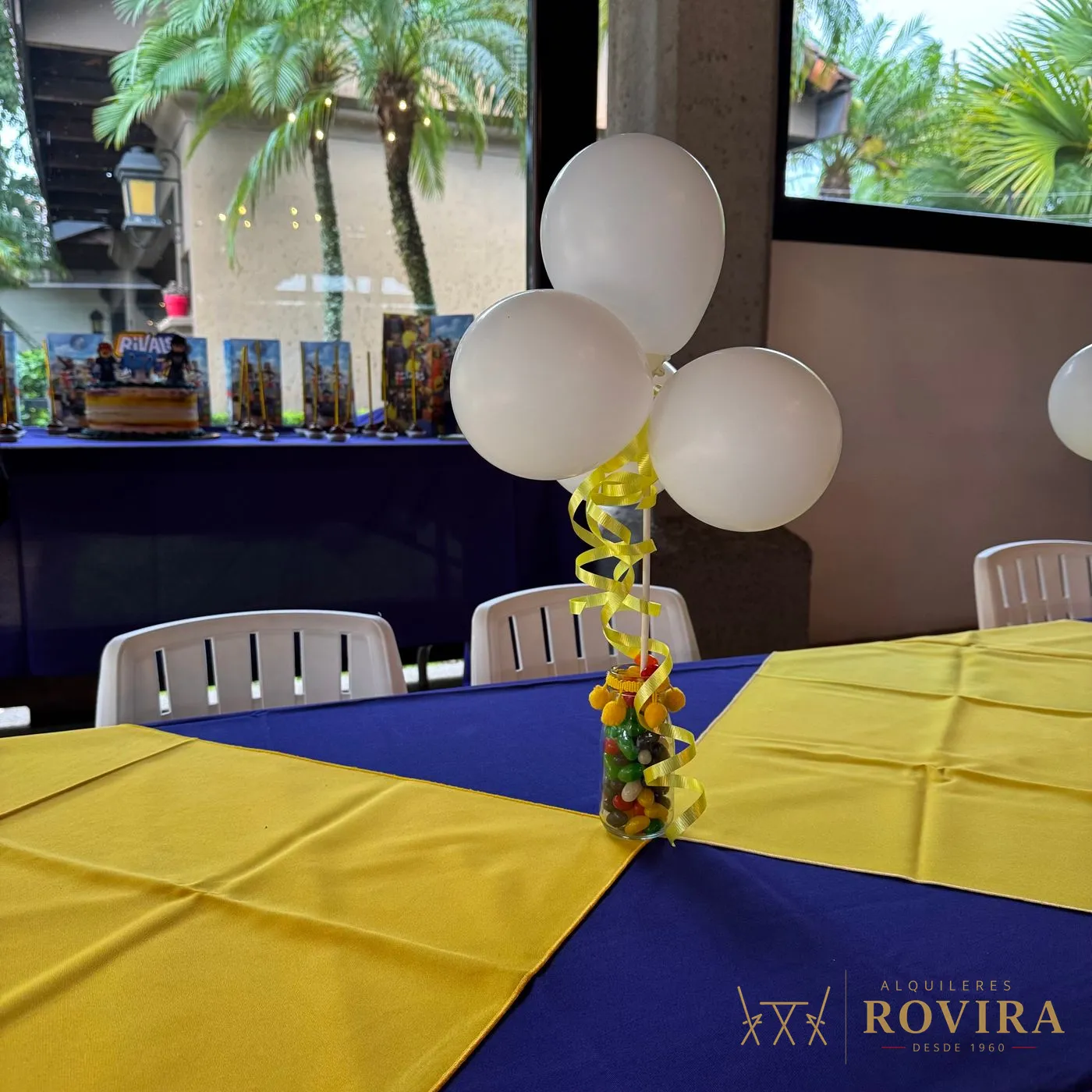 Centro de mesa infantil con frasco de dulces y globos blancos sobre mantel morado y camino amarillo