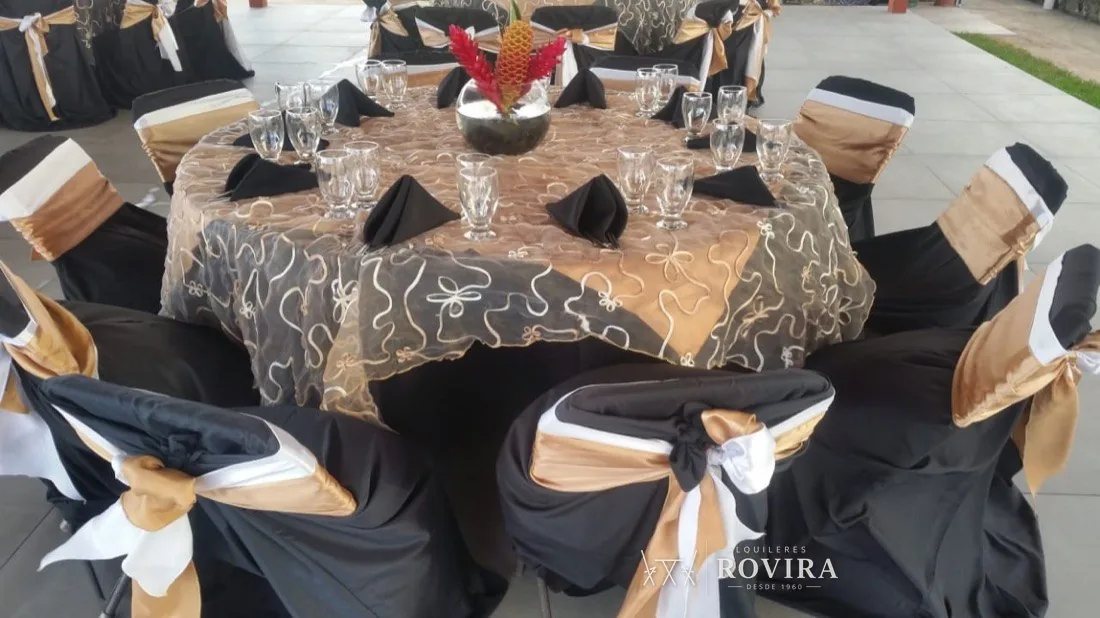 Mesa redonda con mantel dorado y negro estampado y silla negra con lazo tricolor dorado, blanco y negro