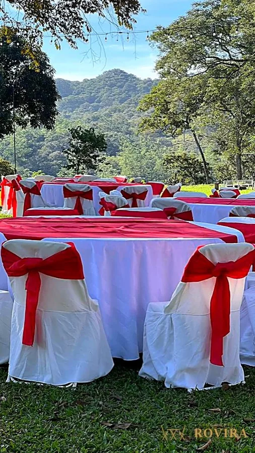 Mesas redondas con mantel blanco y lazos rojos en jardín con vista a la montaña