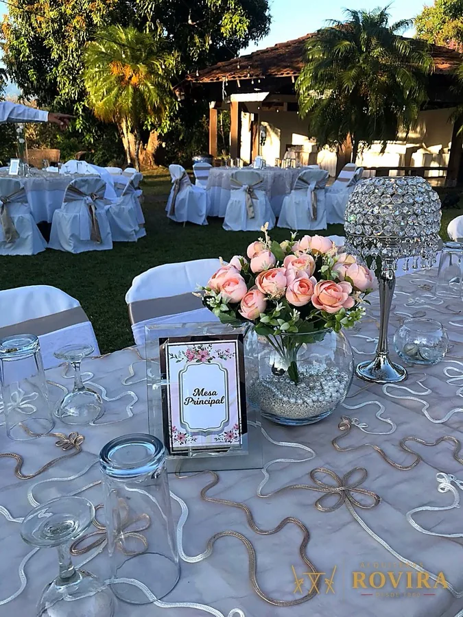 Sillas blancas y centro floral rosado en jardín, con número de mesa