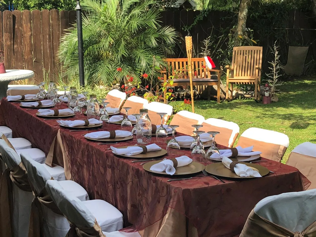 Mesa larga vestida en vino burgundy con lazos café en las sillas, jardín tropical con muro de madera y flores rojas
