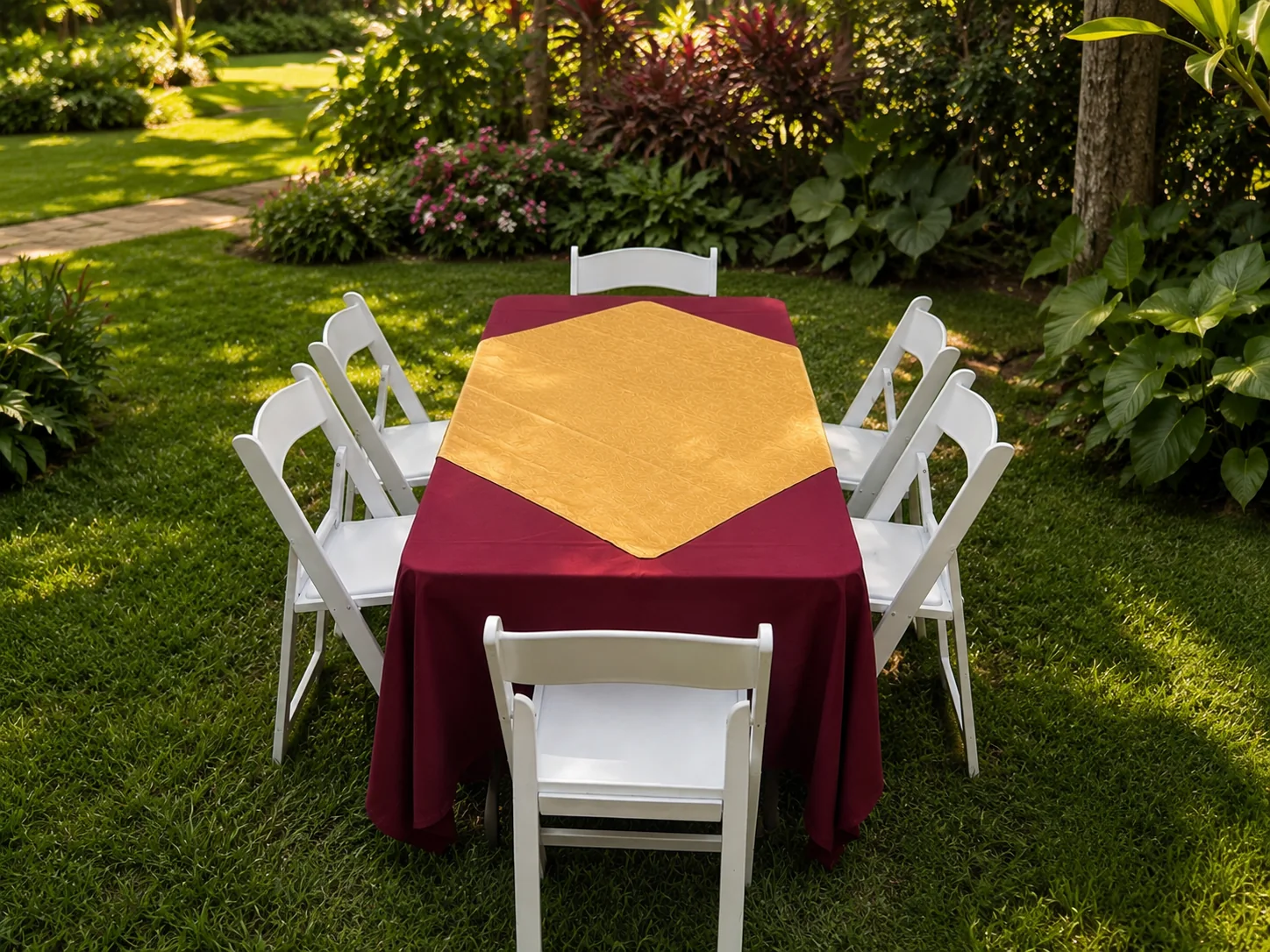 Sillas plegables blancas de Alquileres Rovira alrededor de una mesa rectangular vestida en vino con camino dorado, en un jardín