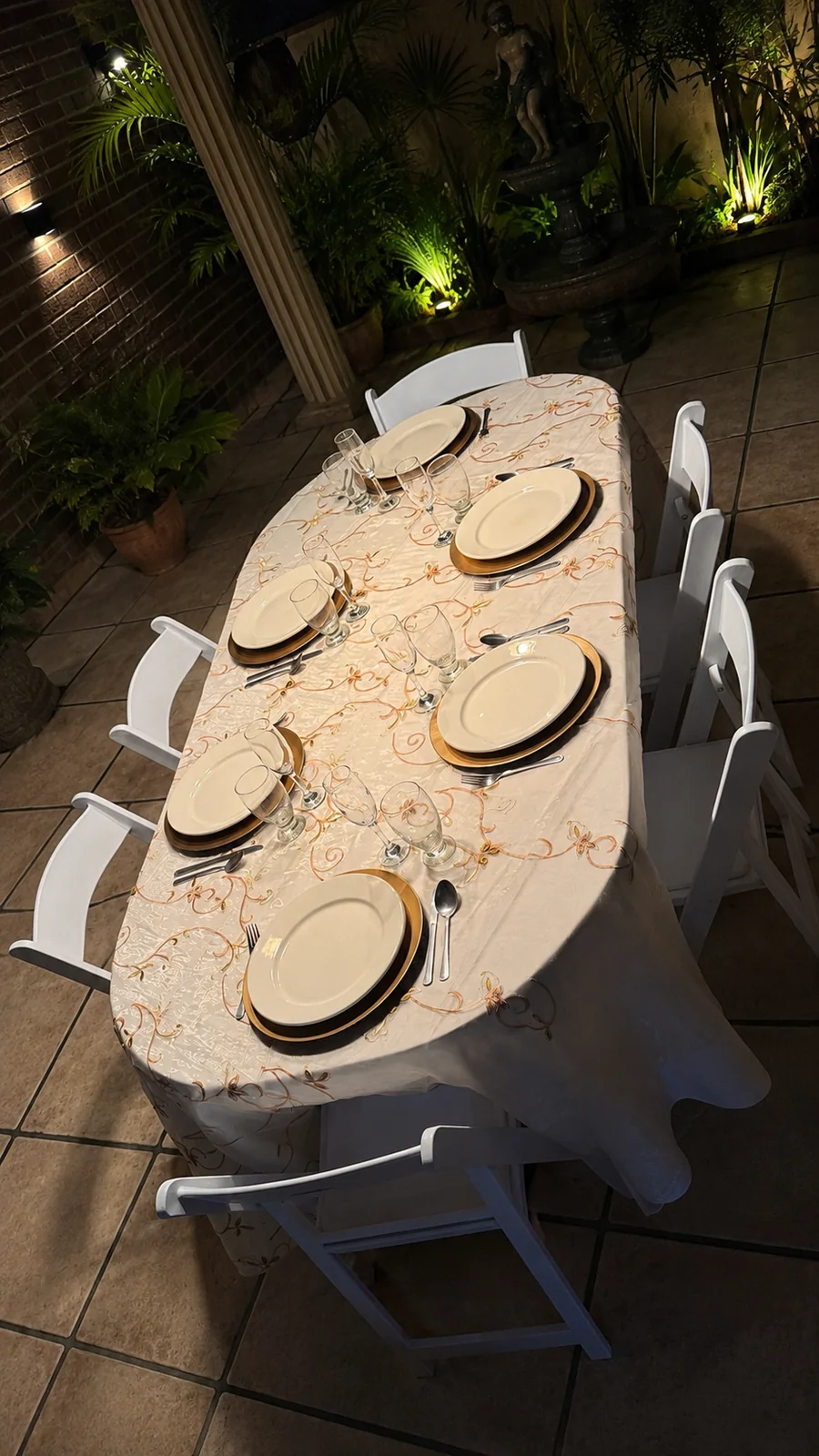 Mesa ovalada montada de noche con platos base dorados, cristalería y sillas blancas de Alquileres Rovira en un patio iluminado con fuente